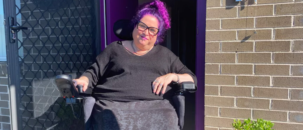 A woman with purple hair, sitting in a wheelchair, in front of her brick house.